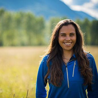 A smiling young woman confidently stands in a field on a sunny day.