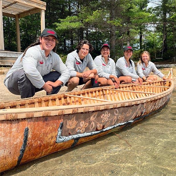 Cinq jeunes Autochtones accroupis à côté d’un canot d'écorce de bouleau traditionnel. 