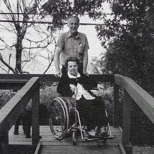 Image en noir et blanc d’une femme en fauteuil roulant en haut d’une rampe d’accès et d’un homme debout derrière elle.