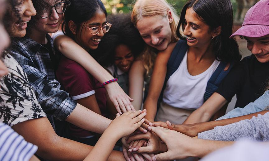 Un groupe d’adolescents souriant gaiement se tiennent en cercle dehors et joignent leurs mains au centre du cercle.