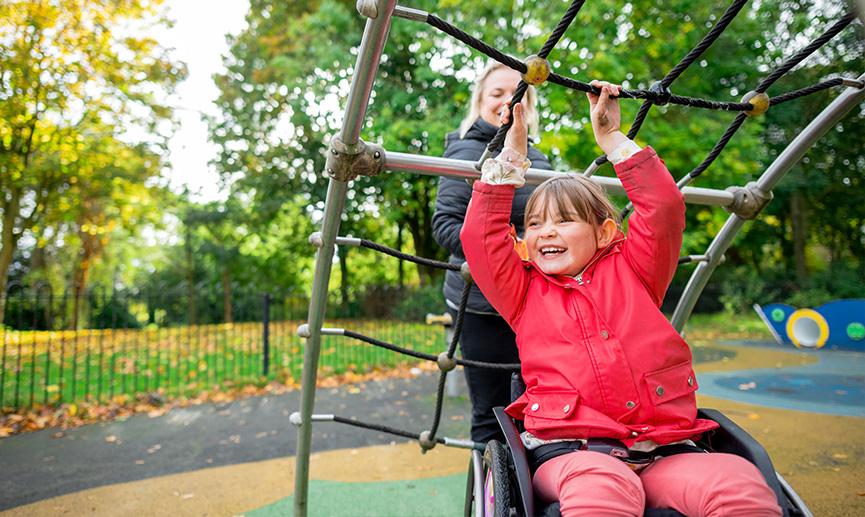 Une jeune personne en fauteuil roulant joue dans un terrain de jeux avec sa maman.