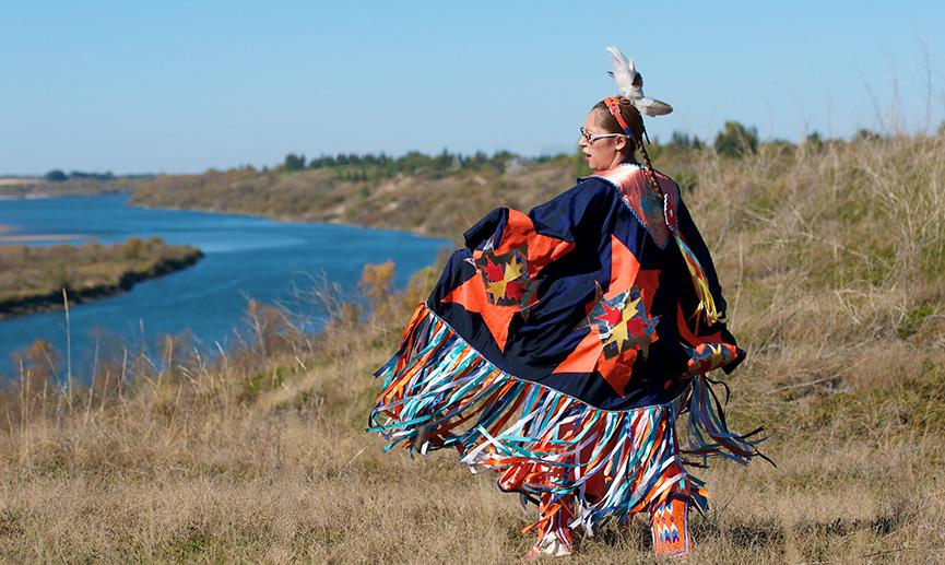 Une femme des Premières Nations exécute une danse libre du châle dans un champ d’herbe avec une rivière à l’arrière-plan.