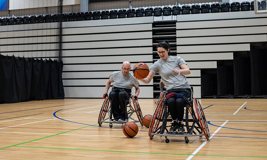 A team of wheelchair basketball players are laughing and dribbling a basketball on a sports court.