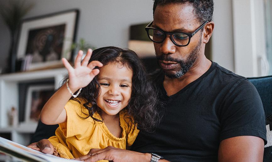 A loving father of African descent sits on the couch in the living room of his home and reads a picture book to his preschool age daughter, who is sitting on his lap smiling. 