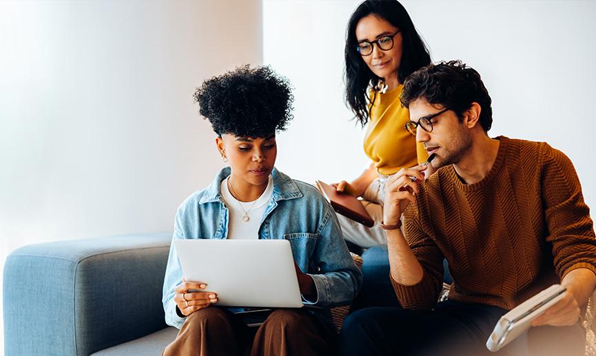 Three colleagues collaborate on a laptop and notes in a relaxed office lounge. Their thoughtful expressions show a calm, engaged atmosphere as they shape their direction for upcoming business work.