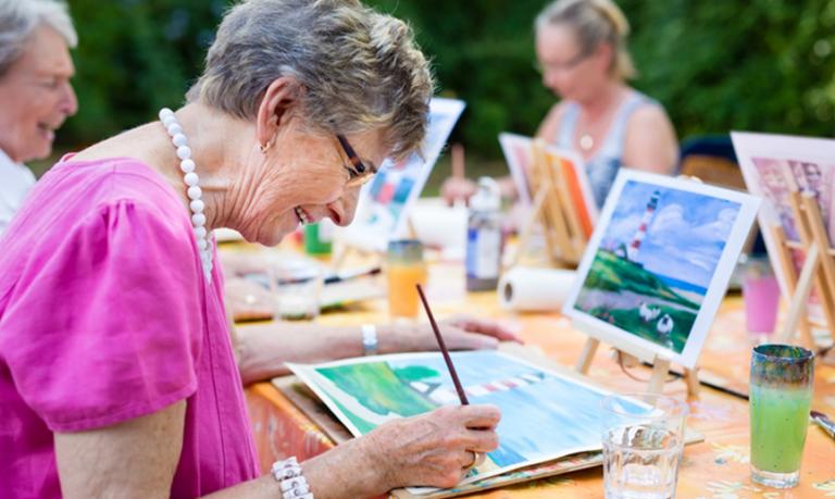 A senior woman paints outside while sitting at a table with a group