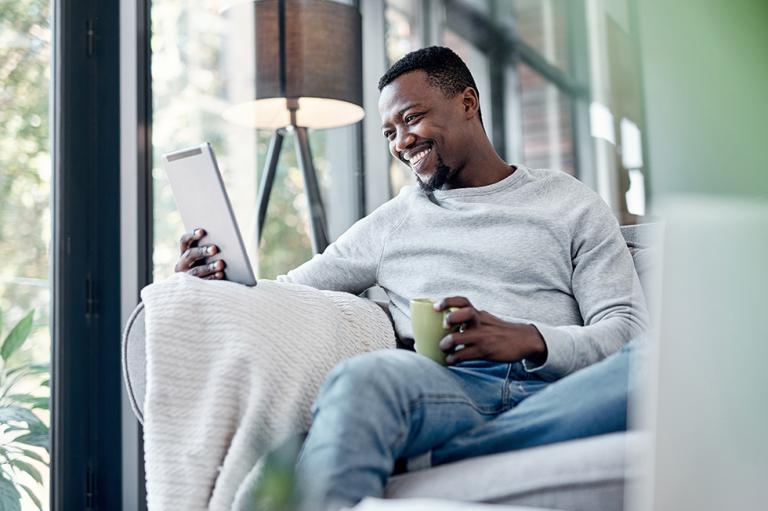 A man using a digital tablet and having coffee on the sofa at home.