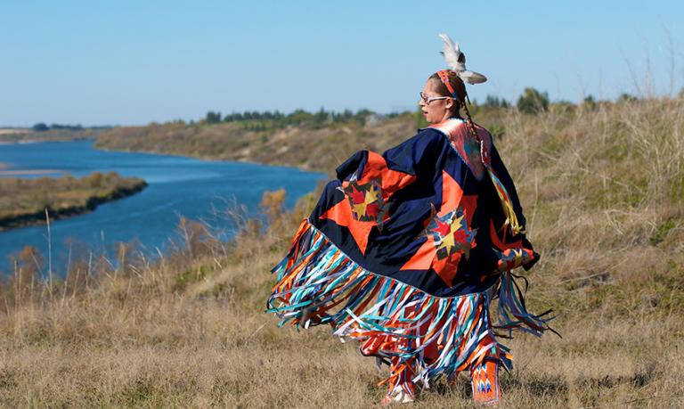 First Nations Woman performs a Fancy Shawl Dance in a grass field with a river background.