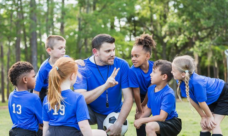 Six multi-ethnic children 6-7 years old playing soccer at summer camp. They are gathered around their coach who is talking strategy.