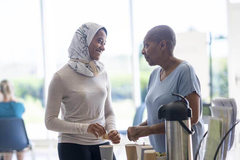 Two people standing at a refreshment table, holding cups and talking in a bright indoor community space.