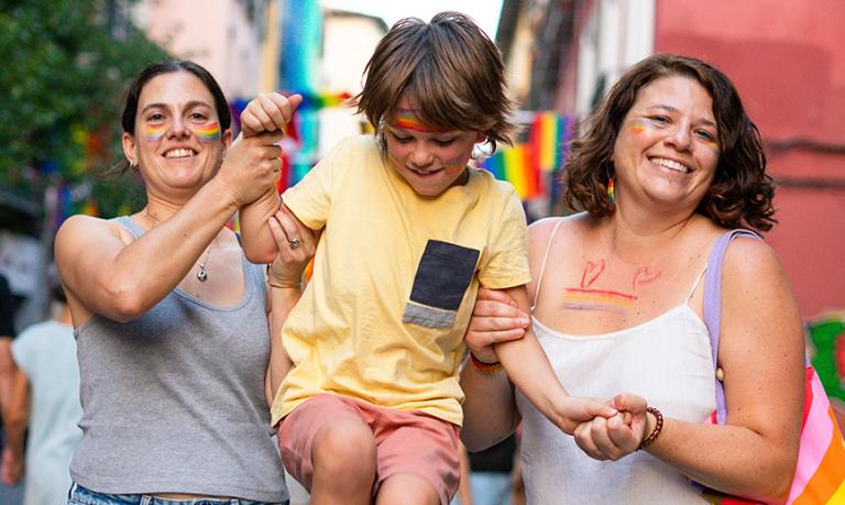 Two smiling mothers are holding their child at a gay pride parade, celebrating with rainbow flags and body paint.